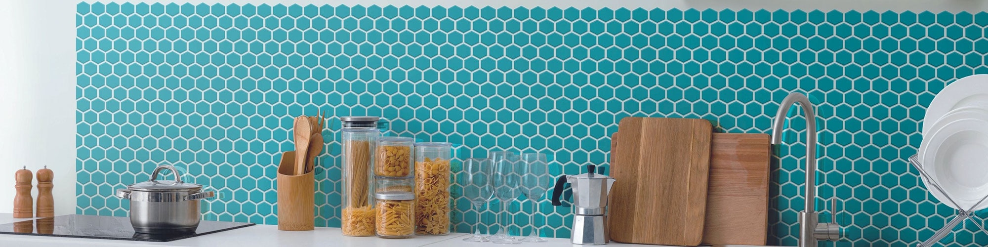 Kitchenette with white quartz countertop, bright teal hexagon mosaic tile backsplash, cutting boards and white plates in a rack.