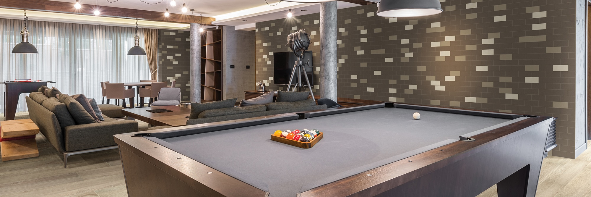 Game room with gray felt billiard table, floor tile that looks like wood, white, tan & brown subway tile, wood beam ceiling, and brown over-stuffed couches.