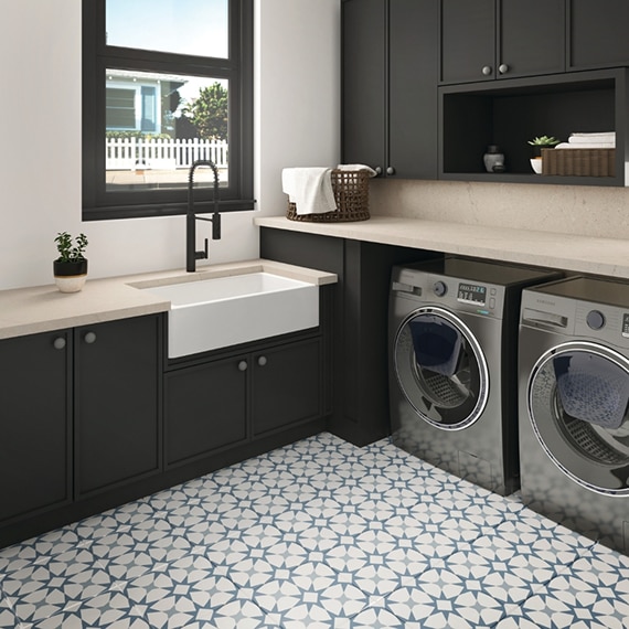 Laundry room with blue & white encaustic floor tile in herringbone pattern, front-loading washer and dryer, brown cabinets, beige quartz counters and backsplash that look like stone.