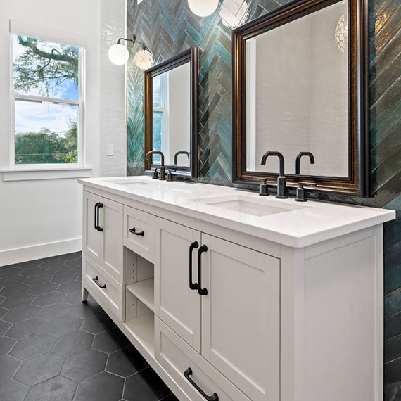 Renovated bathroom with herringbone teal glossy tile backsplash, white quartz countertops, white cabinets, and black hexagon tile flooring.