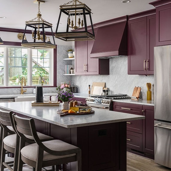 Renovated kitchen with plum-colored cabinets, gray marble backsplash and countertops, stainless steel appliances, and farm sink in front of three windows.