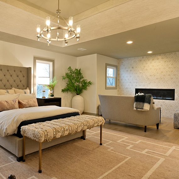 Bedroom with cushioned headboard, nightstands, and wall-mounted fireplace with tan marble and limestone rotating hexagon mosaic wall tile.