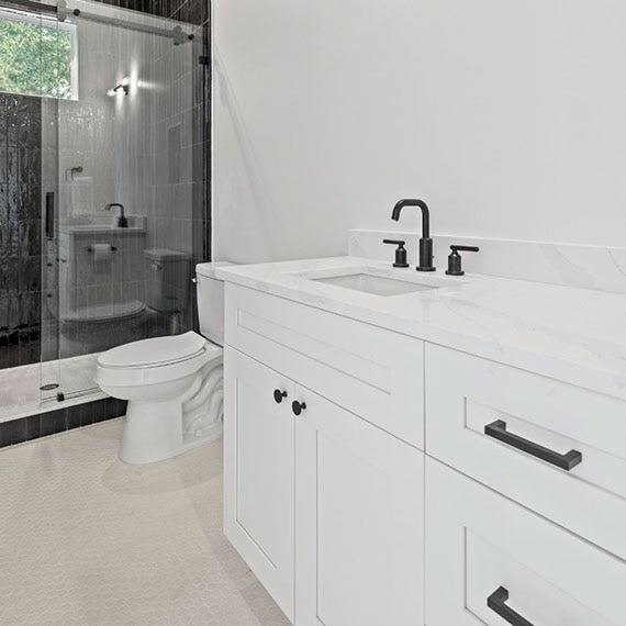 Renovated bathroom with white quartz countertops, white cabinets with black fixtures, black glossy shower wall tile, and white hexagon mosaic floor tile.