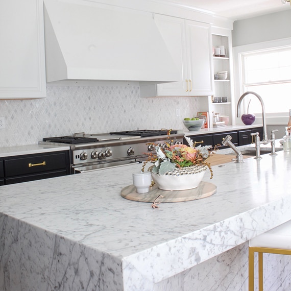 Kitchen with marble mosaic backsplash, white quartz countertops, marble waterfall island with sink, white and black lower cabinets, and stainless steel gas stove/oven.