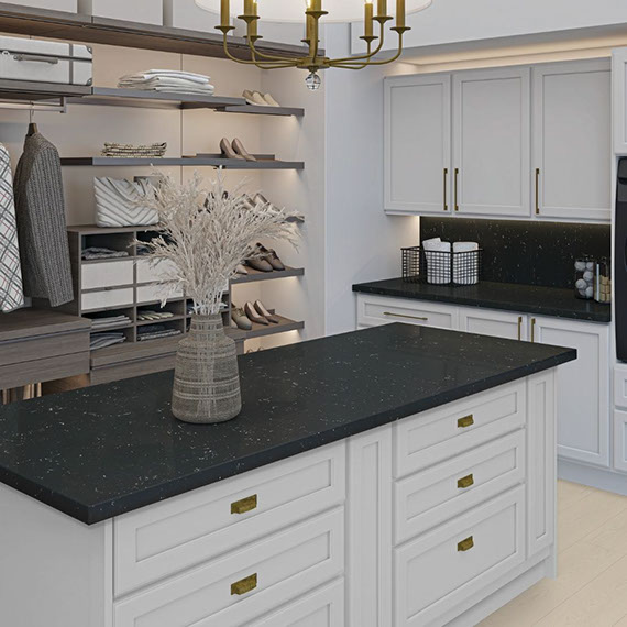 Large walk-in closet with white built-in cabinets and chest of drawers with black quartz countertops, clothing on hangers, floating shelves with shoes & accessories.