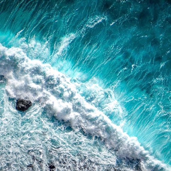 Bird's eye view of aqua ocean waves crashing over rocks at the shoreline.