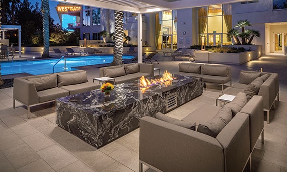 Condominium pool with blue tile, deck of large format taupe tile that looks like stone, palm trees, covered patio with firepit surrounded by tan couches.
