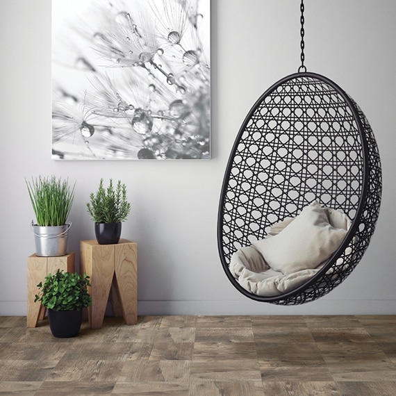Hanging wicker chair, 10x10 wood look tile flooring with potted plants on wood pedestals and large black & white print on the wall.