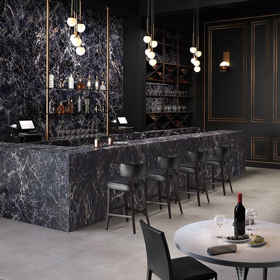 Restaurant bar with black with white veining porcelain slab on the wall, bar top and front, brass and globe pendant lighting, gray stone look flooring tiles.