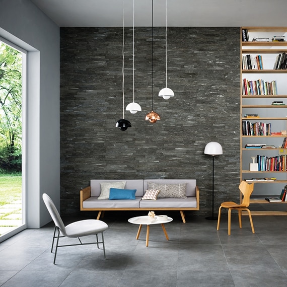 Living room with gray floor tile that looks like stone, dark gray wall tile that looks like stacked stone, wood bookshelves, and pendant lights over a white couch.
