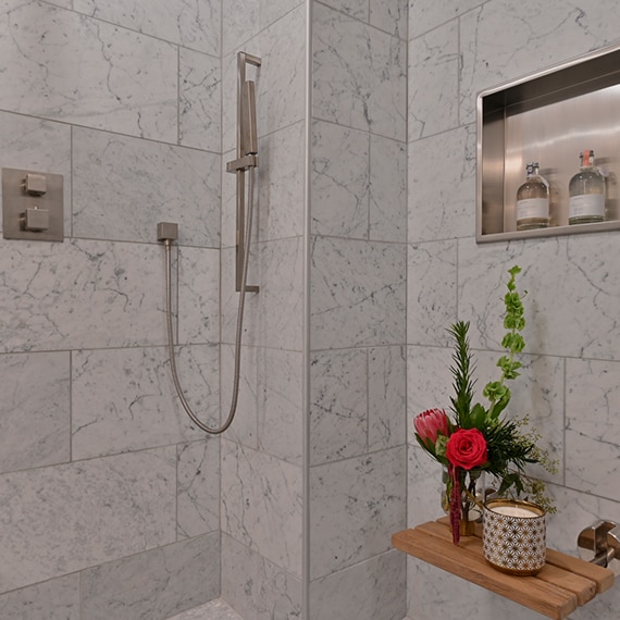 Wet room with white & gray vein marble wall tile, stainless steel shower niche, and floating shelf holding bouquet of red flowers.
