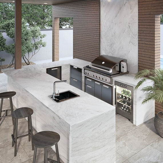Outdoor kitchen with gray stone-look floor tile, backsplash, countertop and island of white quartzite with gray striations, stainless steel grill, wine refrigerator, and wood patio cover.