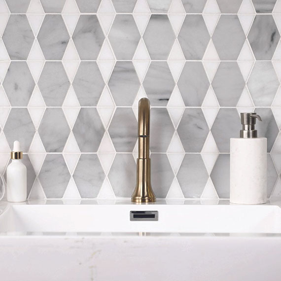 Closeup of bathroom backsplash of pre-cut gray marble and white marble mosaic tile, white stone sick with burnished brass faucet, and white soap dispenser.