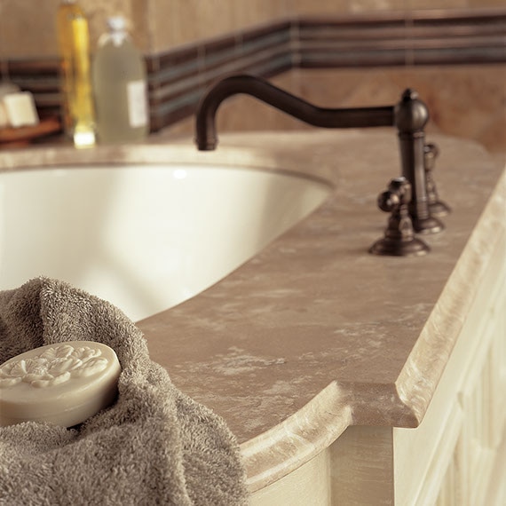 Free-standing bathtub with tan travertine surround, rubbed bronzed faucet, and decorative bar soap on gray towel.