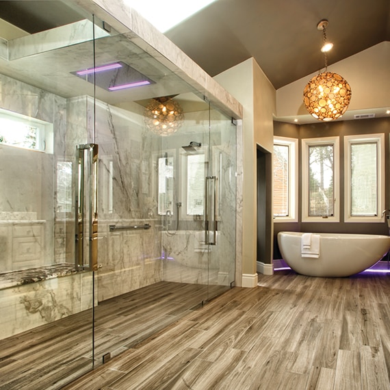 Bathroom with tile floor that looks like wood, large walk-in shower with porcelain slab walls that look like marble, and freestanding tub with globe chandelier.