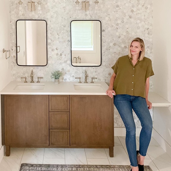 Instagram influencer Jennifer Gizzi sitting on her recently renovated bathroom vanity with white & beige marble mosaic backsplash and marble-look white & gray floor tile.