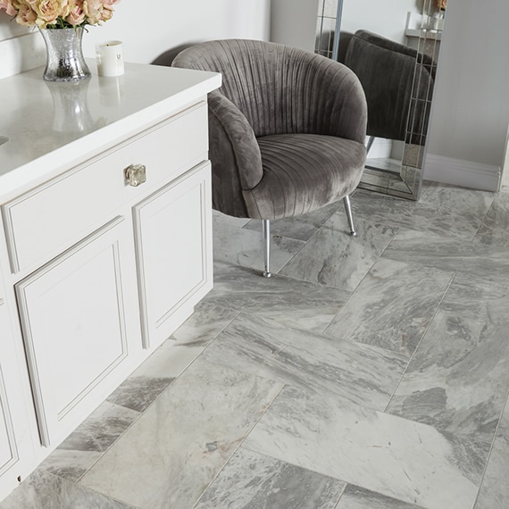 Elegant bathroom with gray marble floor tile, white quartz vanity, white cabinet, and gray velvet chair.