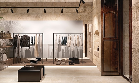 Clothing store with beige floor and wall tile that looks like limestone, racks of women’s clothing, floating wood shelves holding purses and duffle bags. 