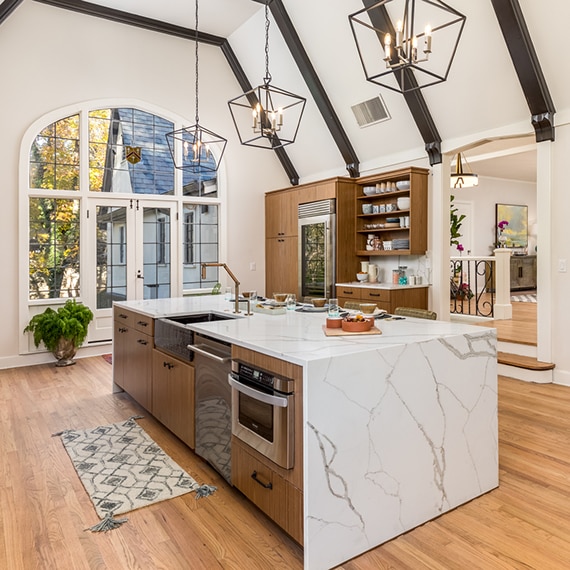 Remodeled kitchen with high vaulted ceiling, large picture windows, white & gray marble look quartz waterfall island and backsplash, wood cabinetry and wood beams..
