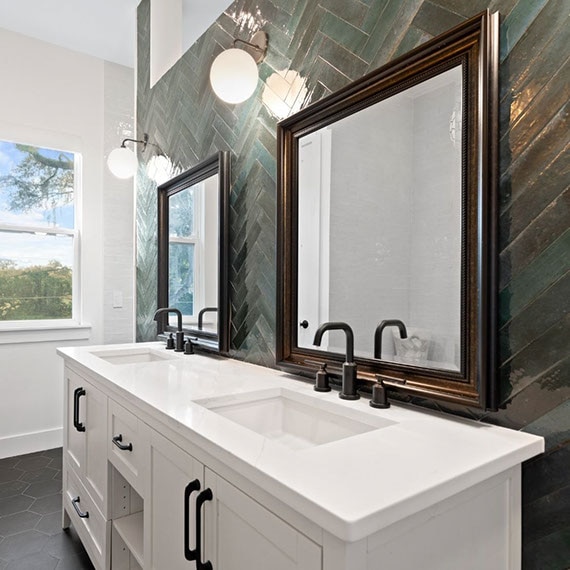 Renovated bathroom with herringbone teal glossy tile backsplash, white quartz countertops, sinks with burnished bronze faucets, and framed mirrors.