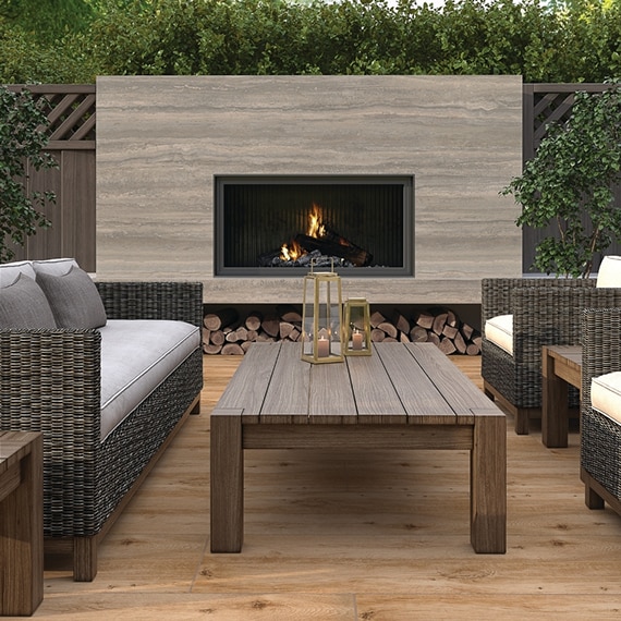 Outdoor patio fireplace with beige porcelain slab surround that looks like travertine,  wicker sofa & chair with gray cushions, and wooden coffee table.