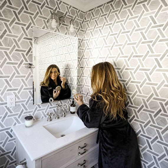 Dolores Catania in her half bath with gray limestone & white marble rotating hexagon mosaic wall tile and white wood vanity with white marble countertop.